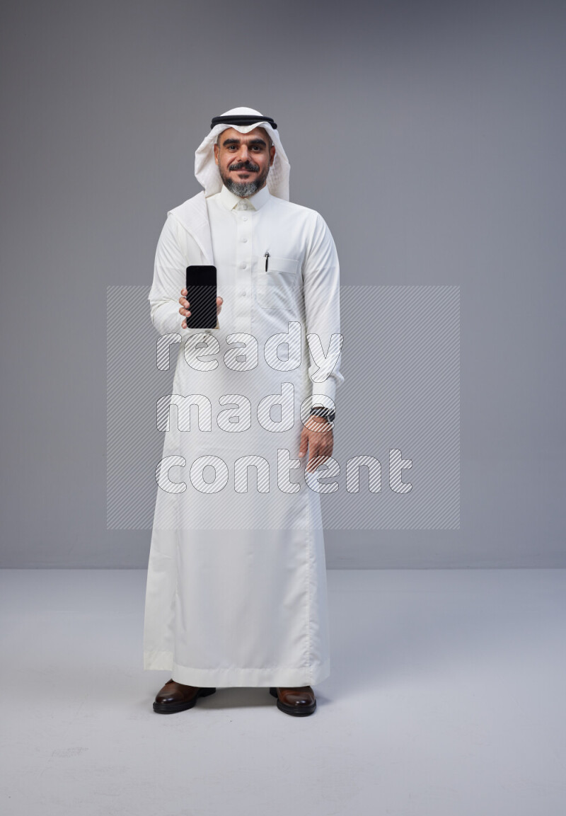 Saudi man Wearing Thob and white Shomag standing showing phone to camera on Gray background