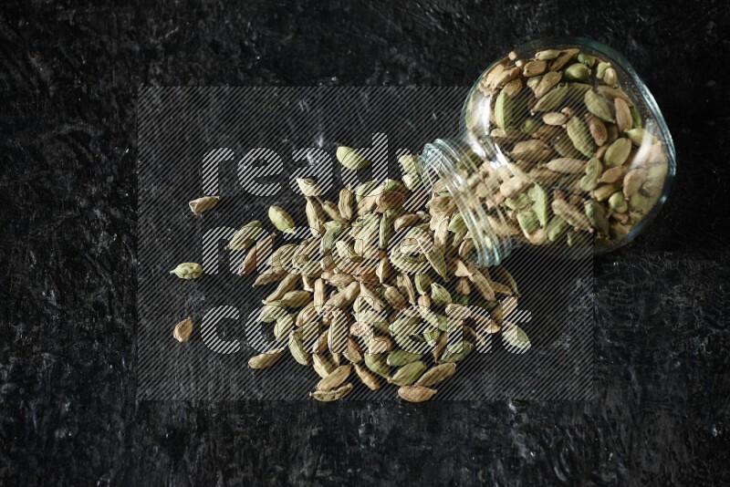 A flipped glass spice jar full of cardamom seeds on textured black flooring