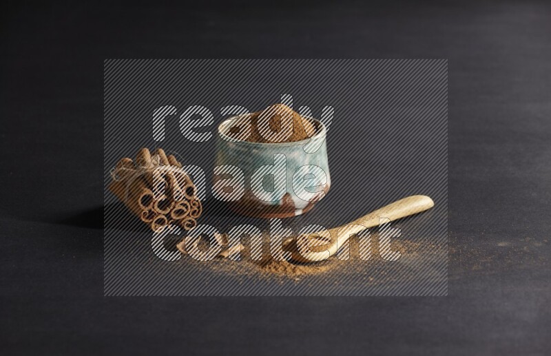 Ceramic bowl full of cinnamon powder and a wooden spoon full of powder with cinnamon sticks stacked and bounded on black background