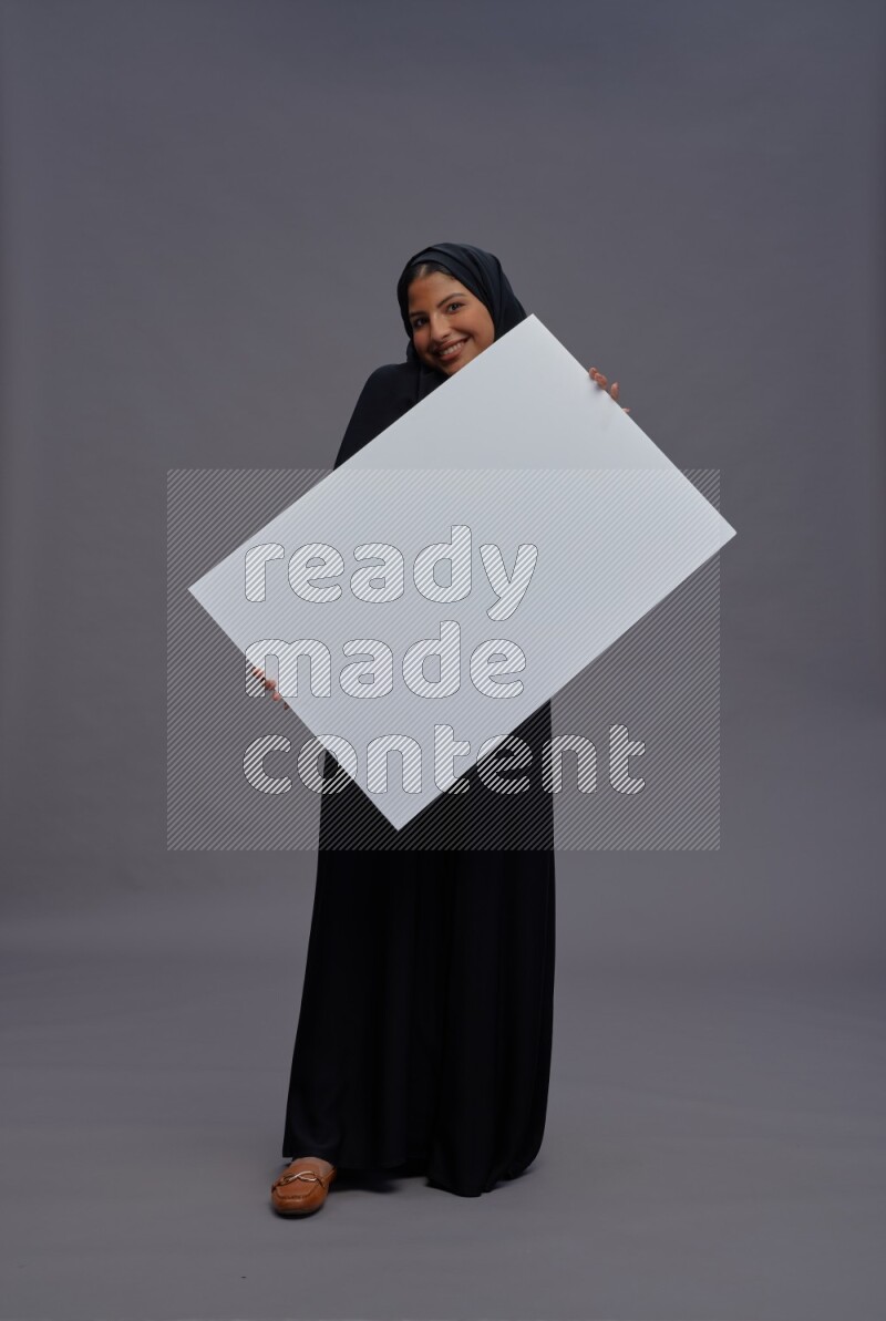 Saudi woman wearing Abaya standing holding white board on gray background