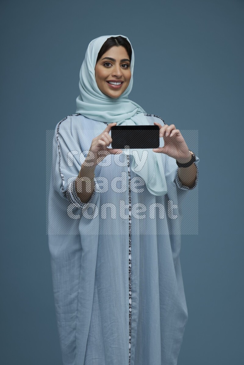 A Saudi woman Showing her phone screen in a blue background wearing a blue Abaya with hijab