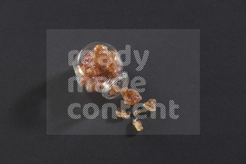 A glass spice jar full of gum arabic and jar is flipped with fallen pieces on black flooring