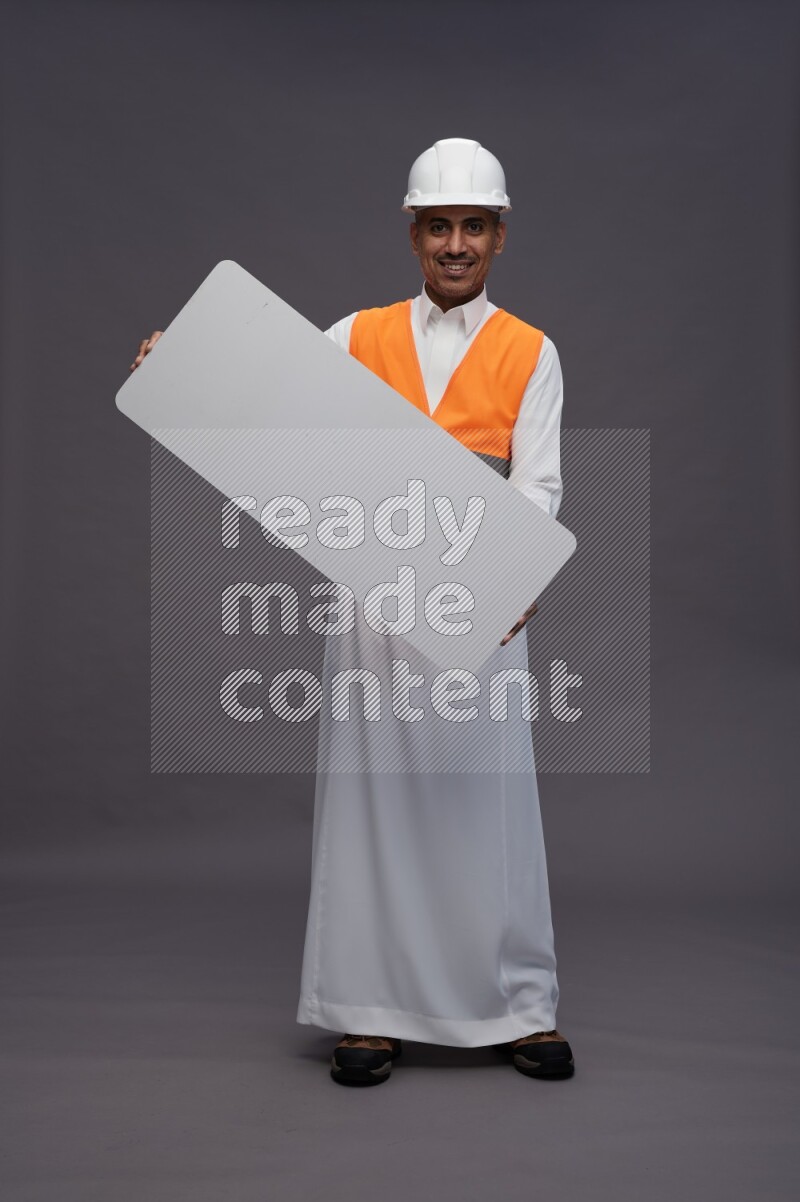 Saudi man wearing thob with engineer vest standing holding board on gray background
