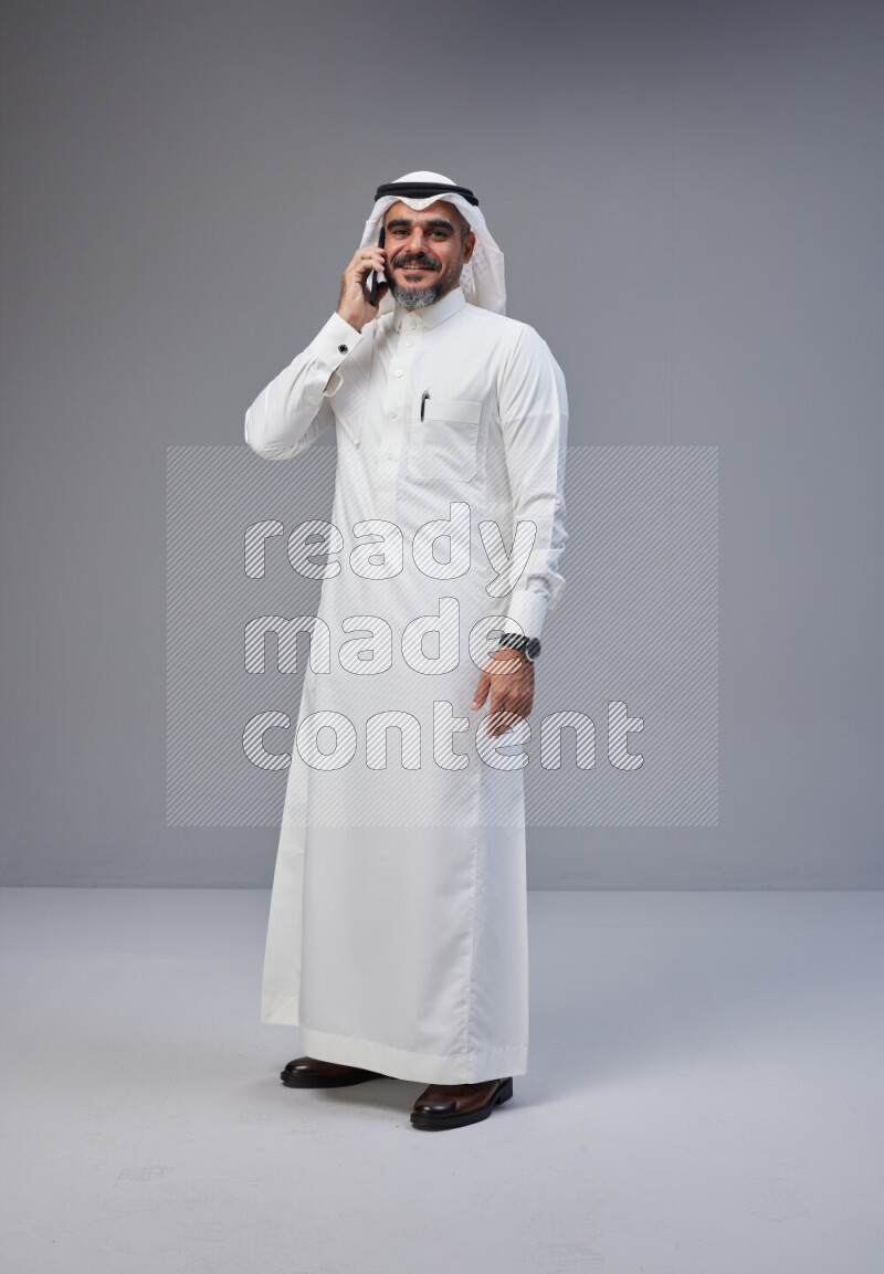 Saudi man Wearing Thob and white Shomag standing talking on phone on Gray background