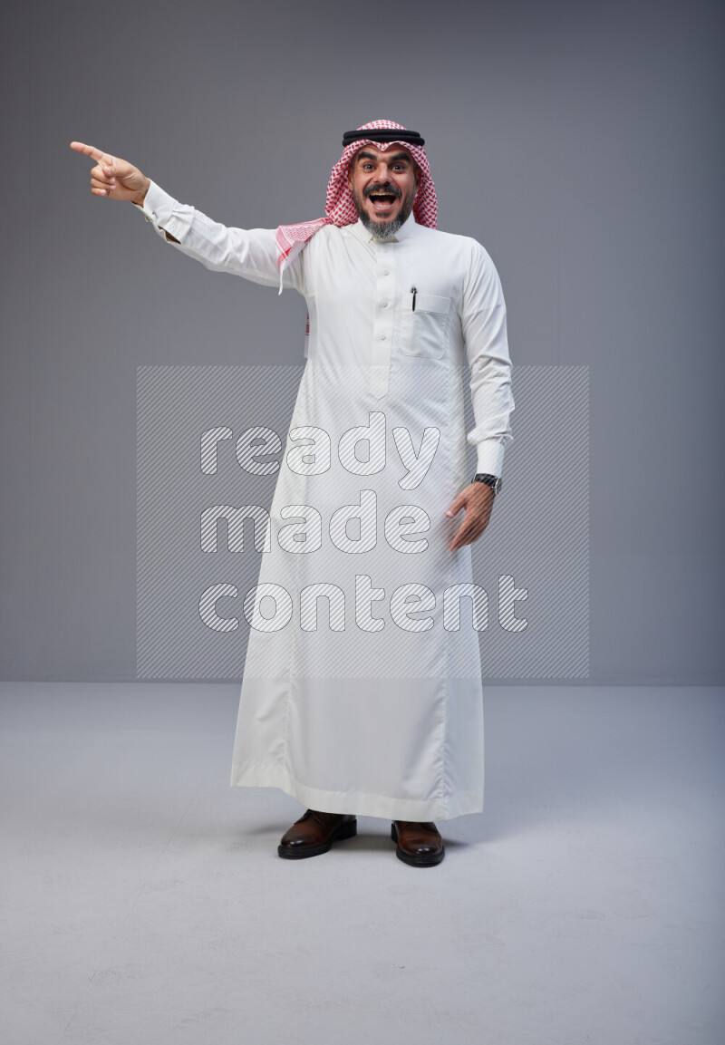 Saudi man Wearing Thob and red white Shomag standing interacting with the camera on Gray background