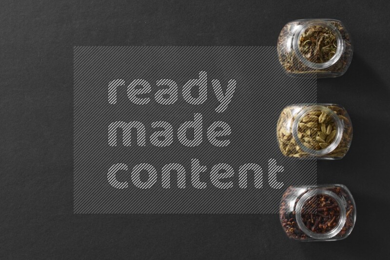 3 glass spice jars full of cloves, cardamom and basil on black flooring