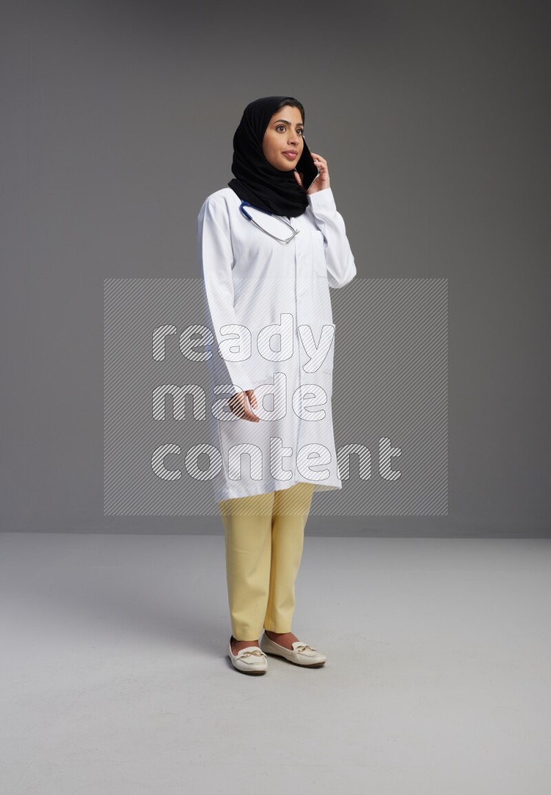 Saudi woman wearing lab coat with stethoscope standing talking on phone on Gray background