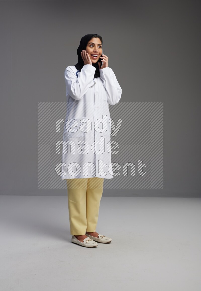 Saudi woman wearing lab coat with stethoscope standing talking on phone on Gray background