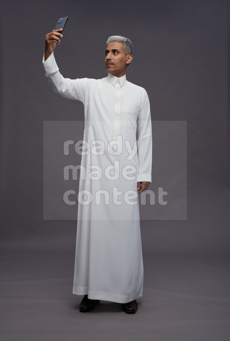 Saudi man wearing thob standing taking selfie on gray background