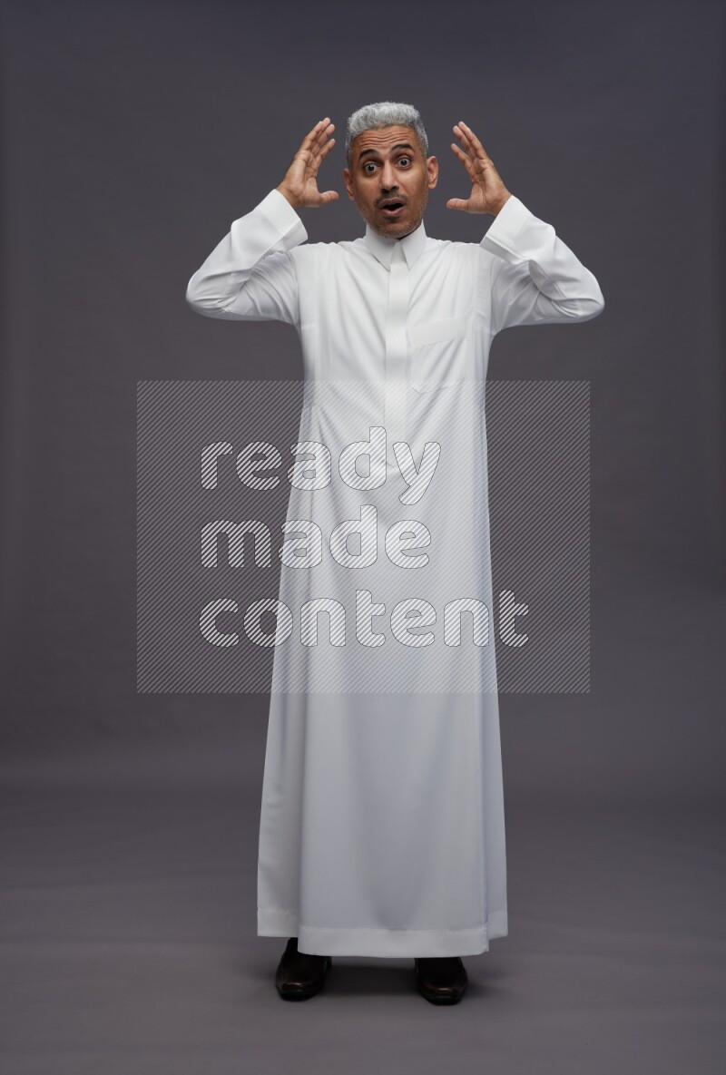 Saudi man wearing thob standing hands behind head on gray background