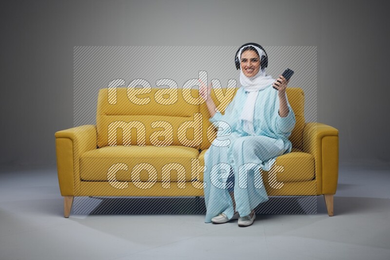 A Saudi woman wearing a light blue Abaya and a white head scarf sitting on a yellow sofa and holding an iPad while wearing headphone eye level on a grey background