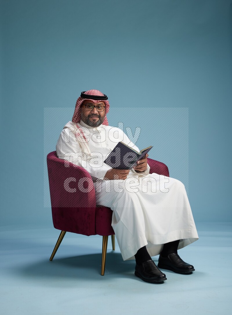 Saudi Man with shimag sitting on chair reading book on blue background