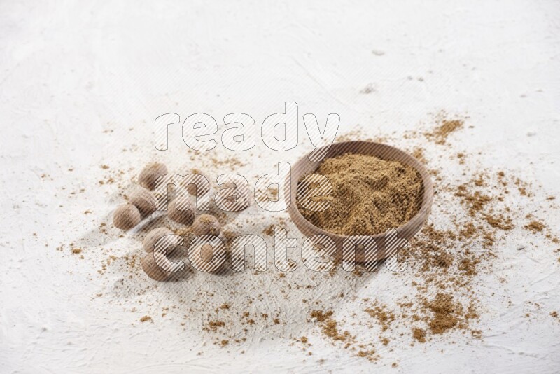 A wooden bowl full of nutmeg powder with whole seeds and sprinkled powder beside it on a textured white flooring