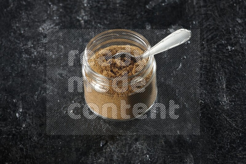 A glass jar and a metal spoon full of cumin powder on a textured black flooring