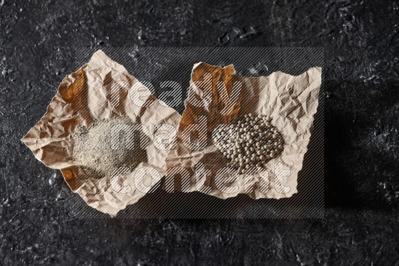 White pepper beads and powder in 2 crumpled paper on textured black flooring