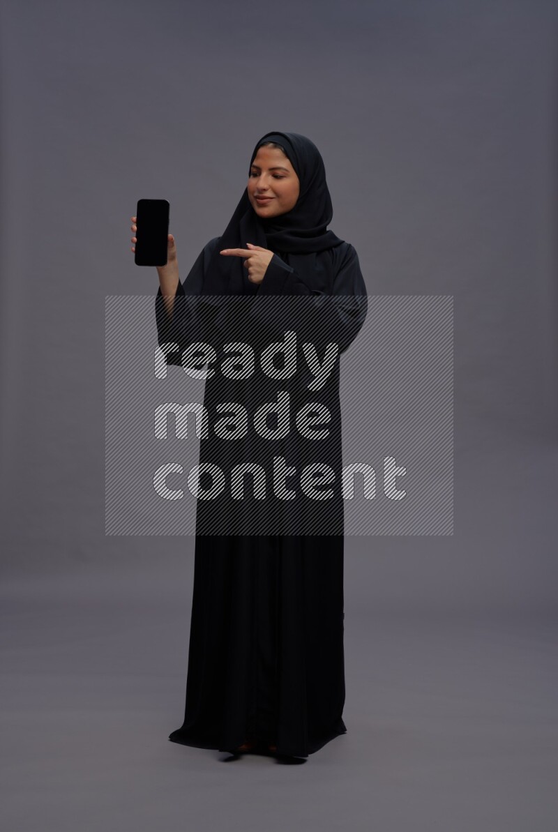 Saudi woman wearing Abaya standing showing phone to camera on gray background