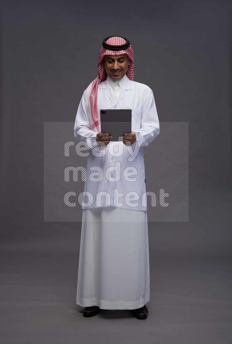 Saudi man wearing thob with lab coat and shomag with pocket employee badge standing working on tablet on gray background