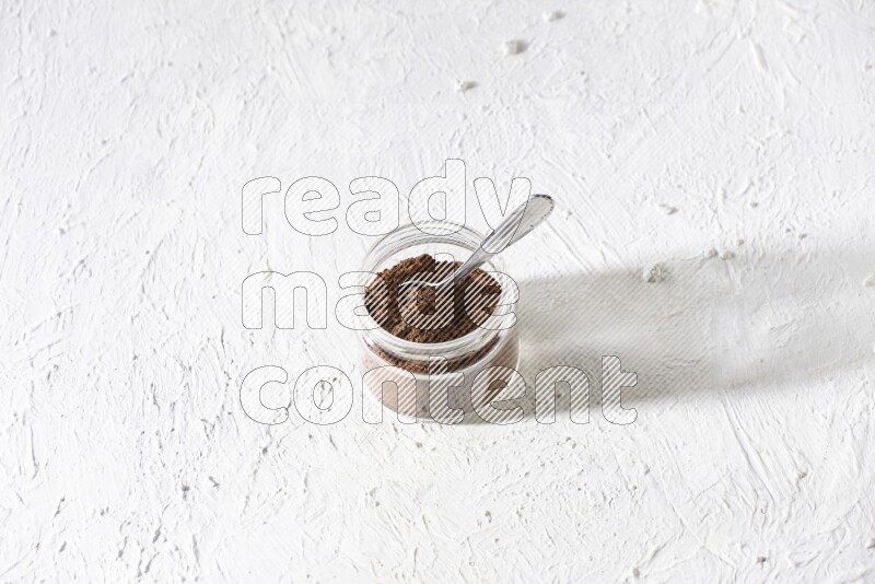 A glass jar full of cloves powder with a metal spoon on a textured white flooring