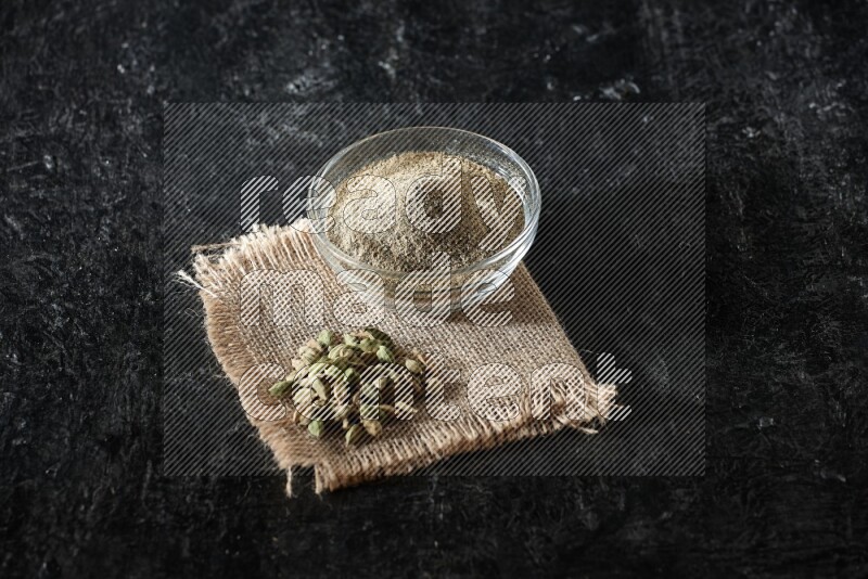 A glass bowl full of cardamom powder with cardamom seeds on a burlap piece on textured black flooring