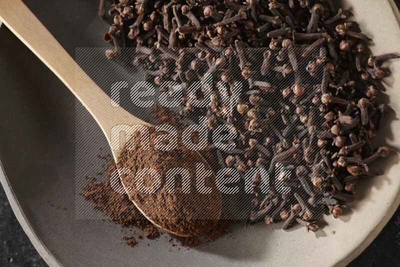 A Pottery plate full of cloves and a wooden spoon full of cloves powder on it on a textured black background