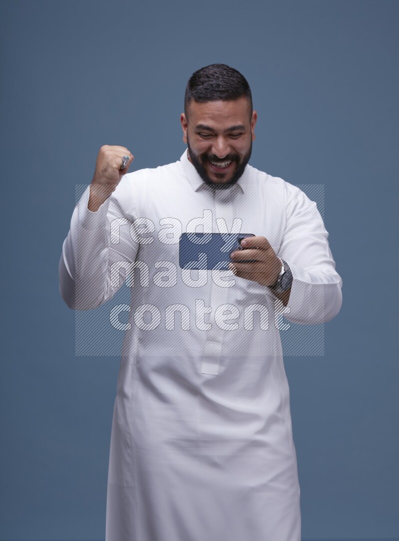 A man Playing a Game on his Smartphone  on Blue Background wearing Saudi Thob