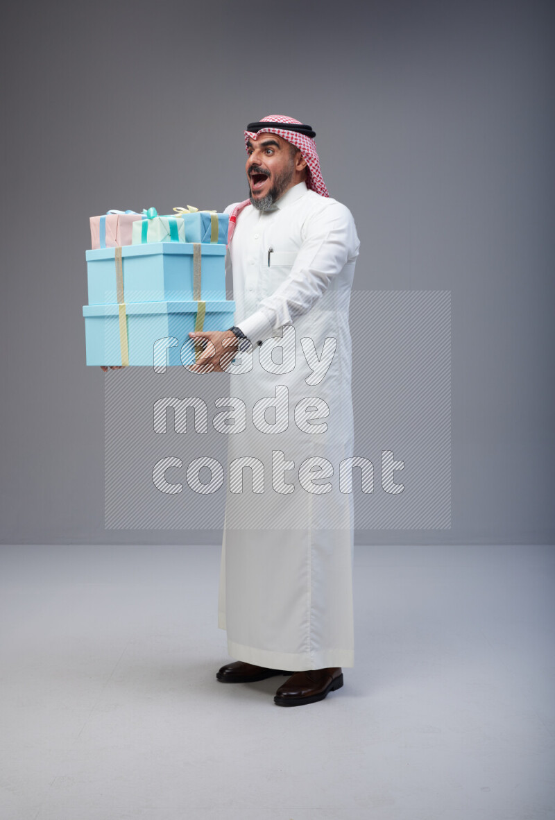 Saudi man Wearing Thob and red Shomag standing holding gift box on Gray background