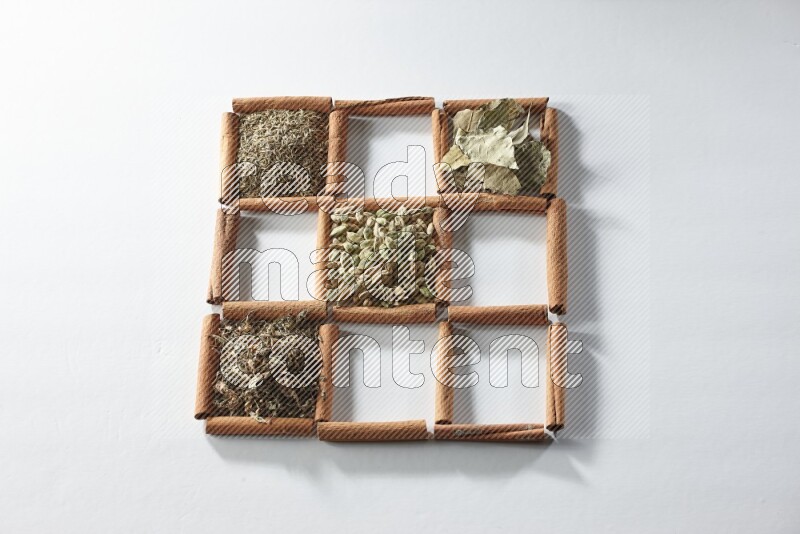 9 مربعات من أعواد القرفة ممتلئة بالهيل في المنتصف محاطة بجوزة الطيب والقرفة وأوراق الغار والقرنفل والكمون والريحان المجفف والزنجبيل المجفف واليانسون النجمي على أرضية بيضاء