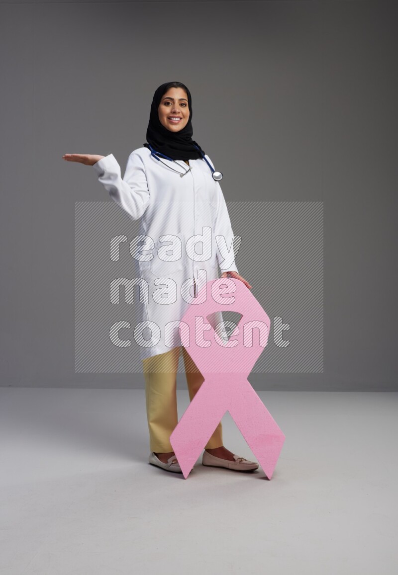 Saudi woman wearing lab coat with stethoscope standing holding awareness ribbon symbol standing on Gray background