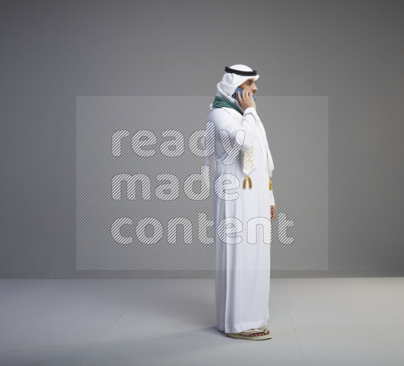 A saudi man standing wearing thob and white shomag with flag scarf on his neck talking on phone on gray background