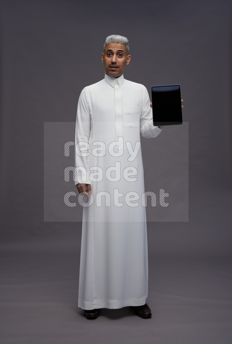 Saudi man wearing thob standing showing tablet to camera on gray background