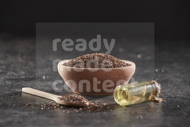 A wooden bowl and spoon full of flaxseeds and a glass bottle of flaxseeds oil on a textured black flooring