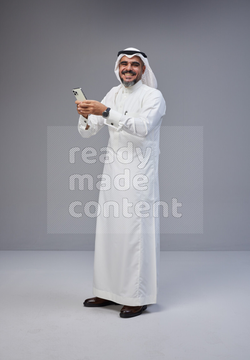 Saudi man Wearing Thob and white Shomag standing texting on phone on Gray background