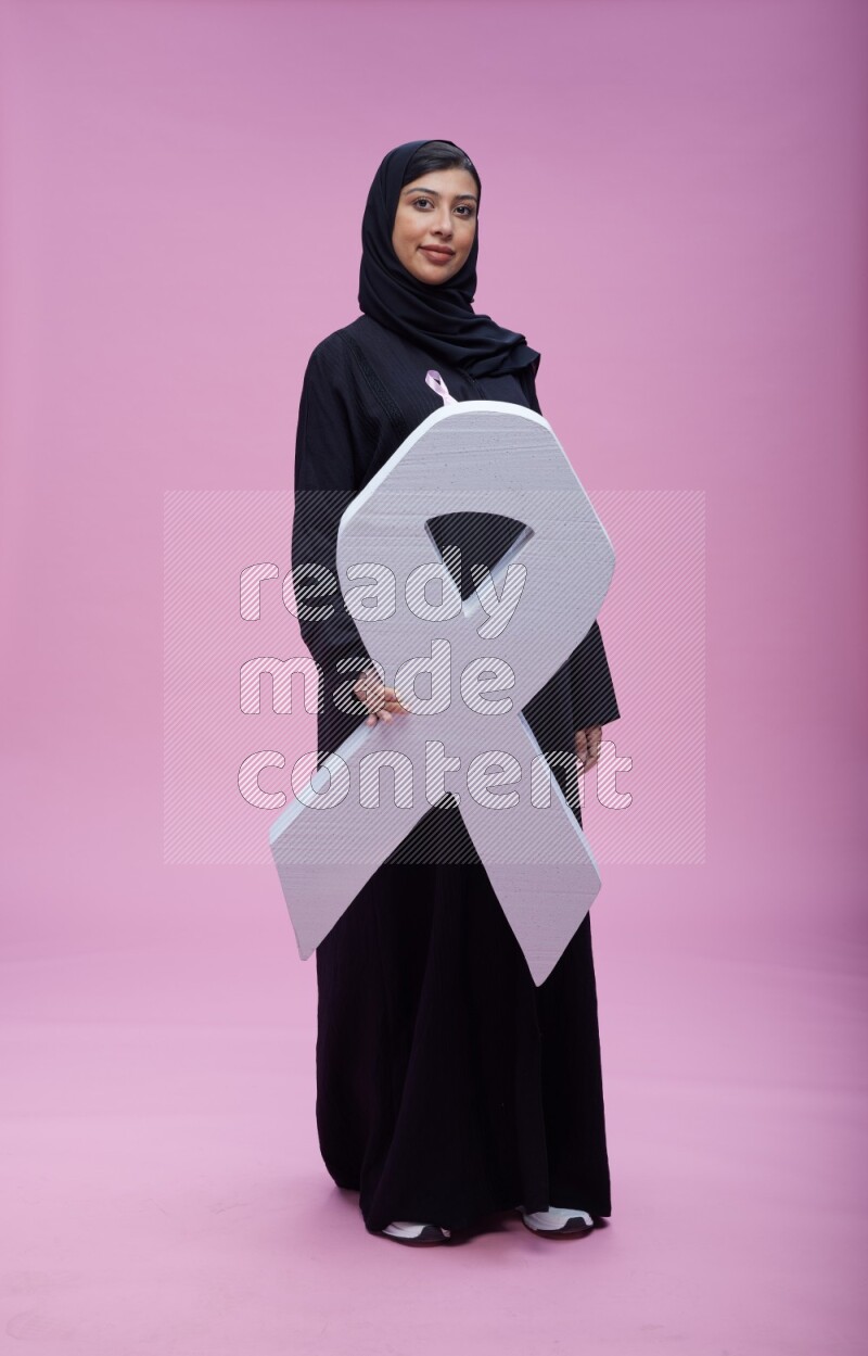 Saudi woman wearing pink ribbon on abaya standing holding awareness ribbon on plain pink background