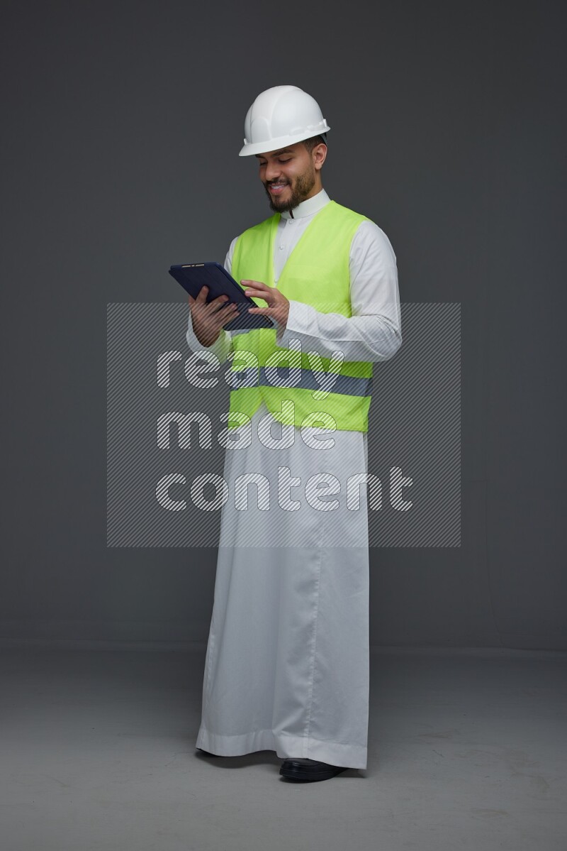 A Saudi man wearing Thobe with a yellow safety vest and white helmet standing and using his tablet different angles eye level on a gray background