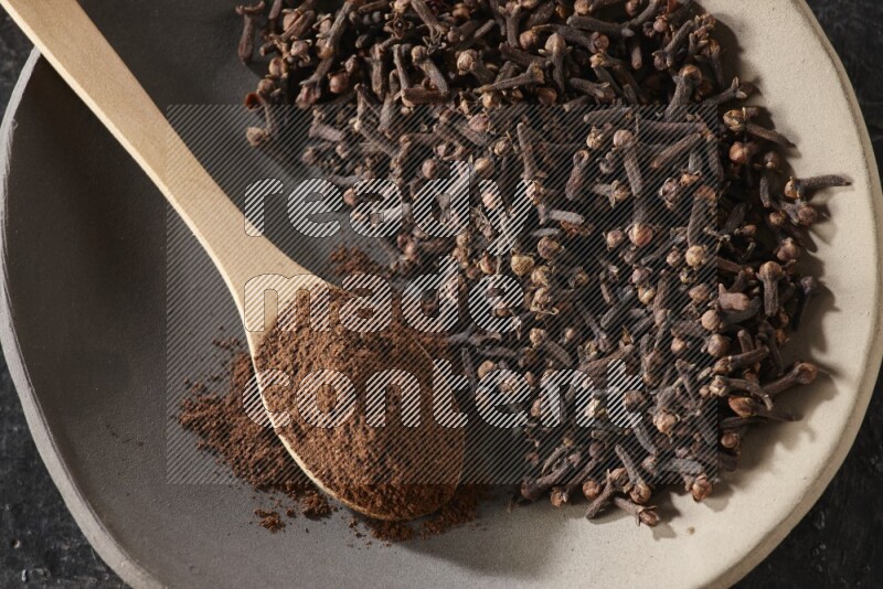 A Pottery plate full of cloves and a wooden spoon full of cloves powder on it on a textured black background
