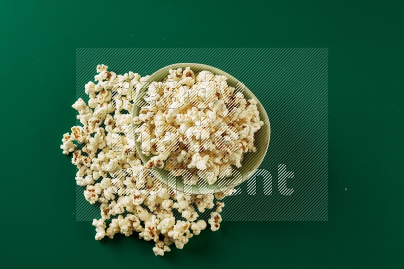A green ceramic bowl full of popcorn with popcorn beside it on a green background in a top view shot