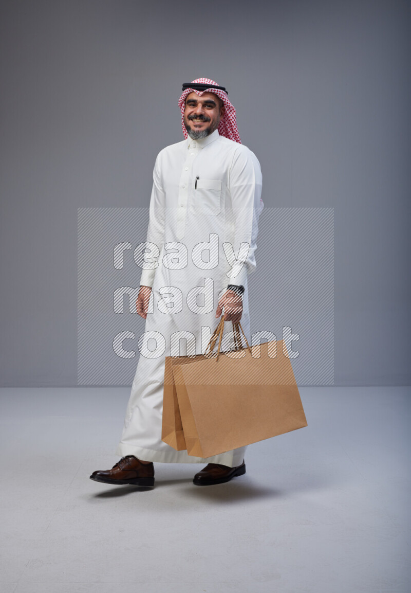 Saudi man Wearing Thob and red Shomag standing holding shopping bag on Gray background