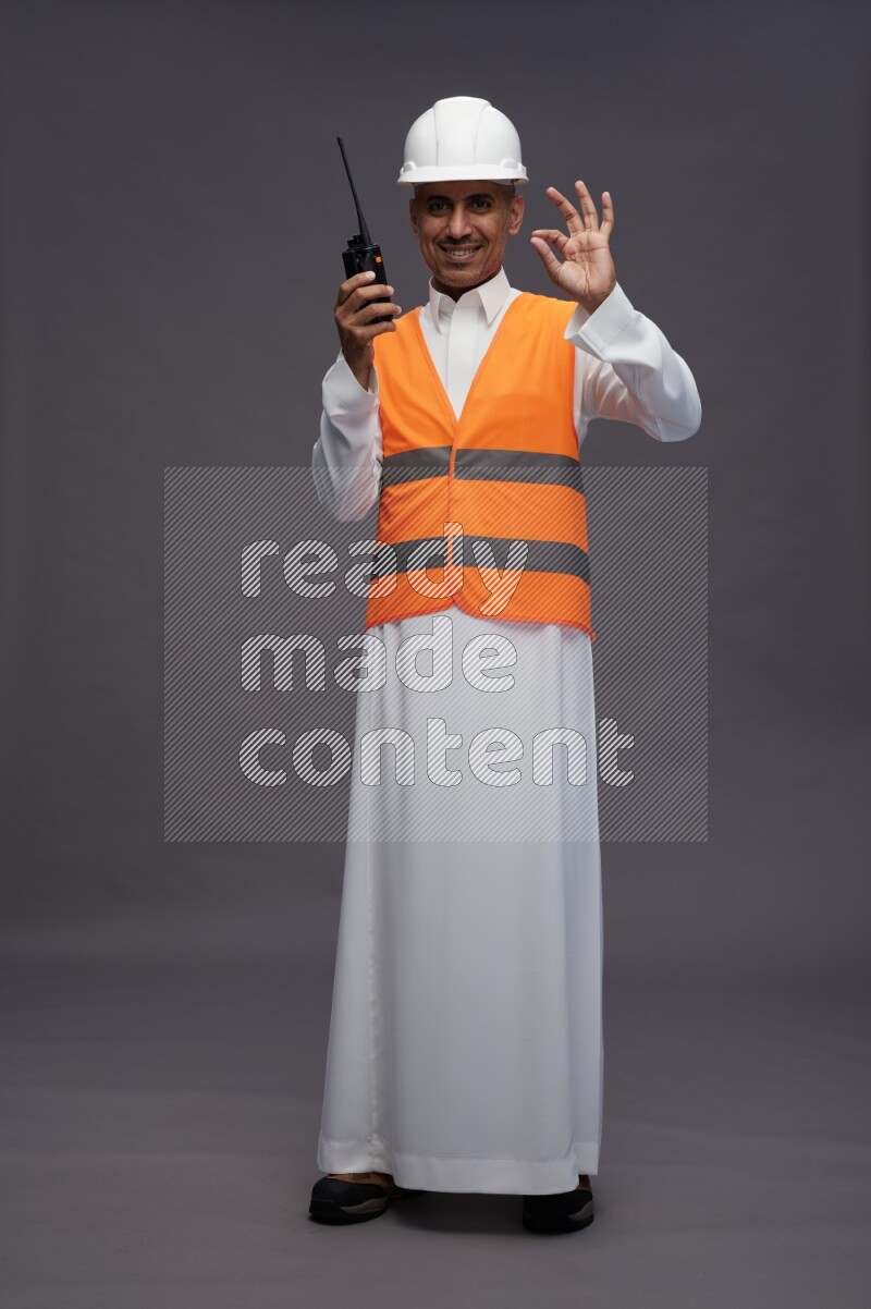 Saudi man wearing thob with engineer vest standing holding walkie-talkie on gray background