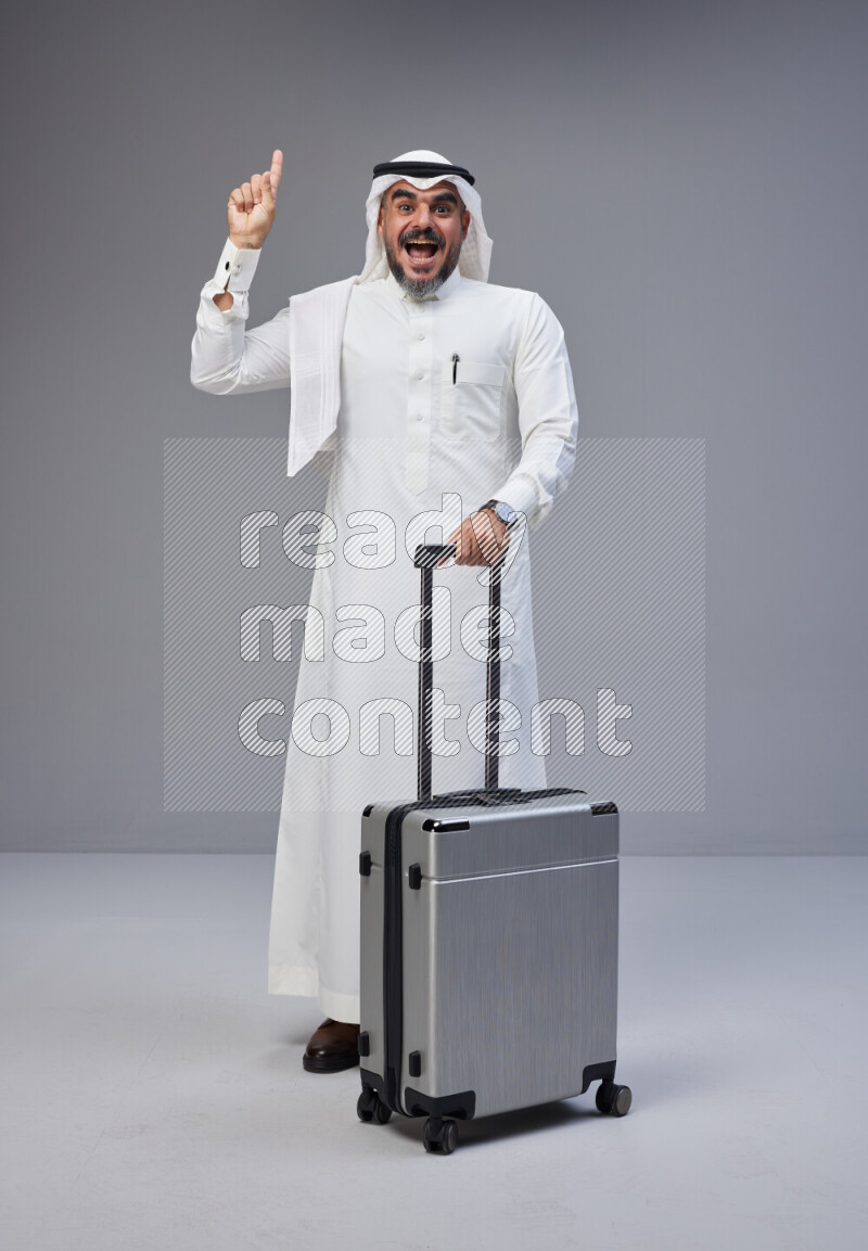 Saudi man wearing Thob and white Shomag standing holding Travel bag on Gray background
