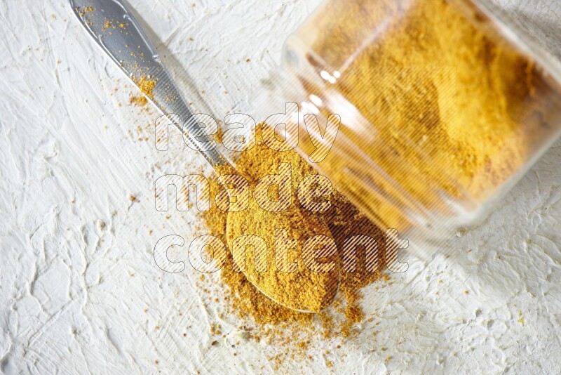 A flipped glass spice jar and a metal spoon full of turmeric powder and powder spilled out of it on textured white flooring