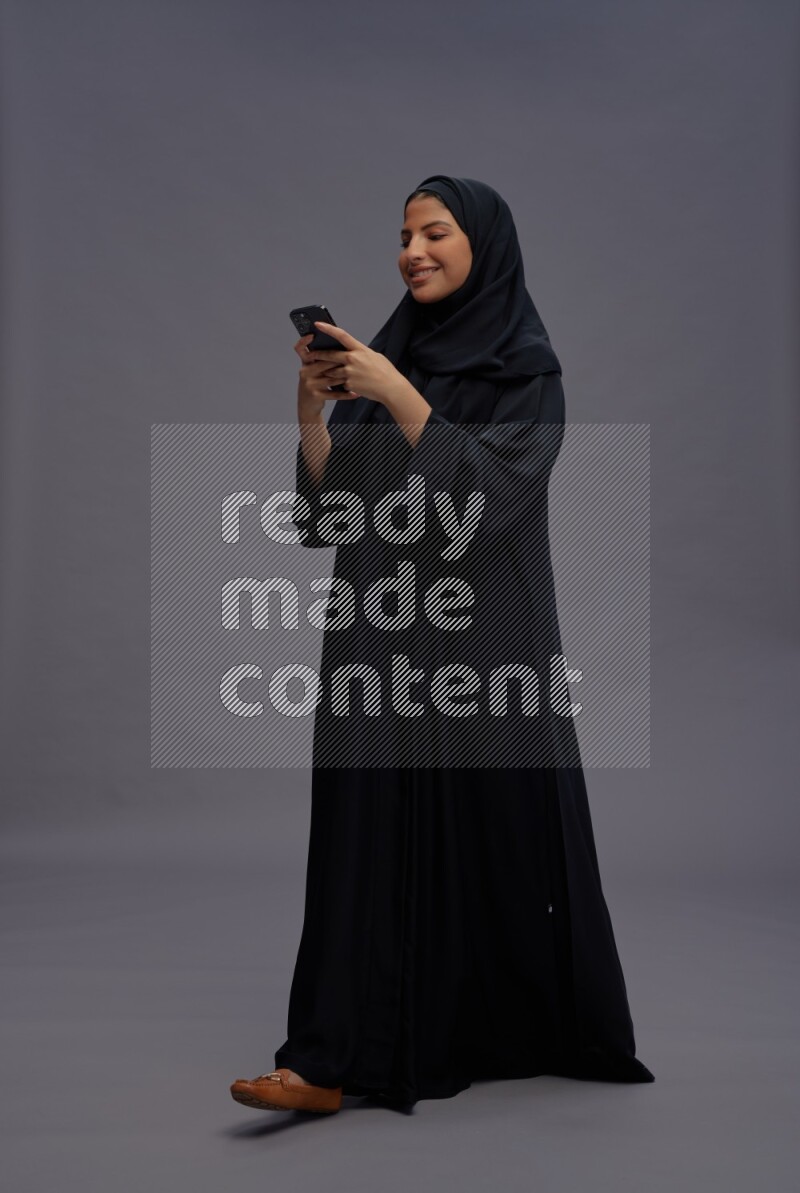 Saudi woman wearing Abaya standing texting on phone on gray background