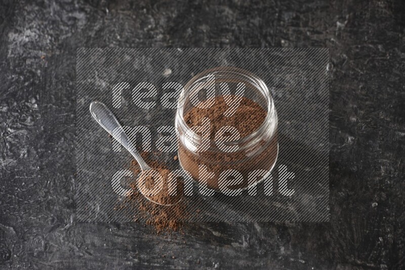 A glass jar full of cloves powder with a metal spoon on a textured black flooring