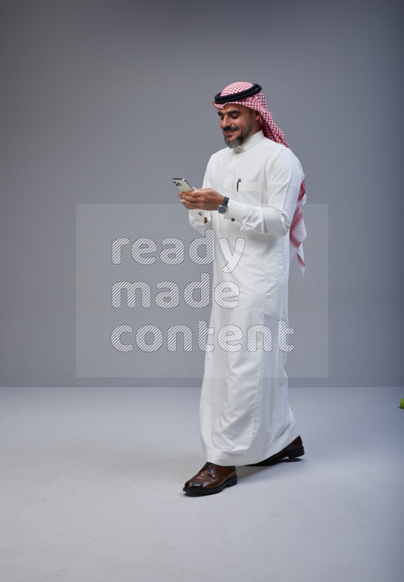 Saudi man Wearing Thob and red Shomag standing texting on phone on Gray background