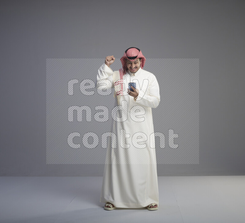 A  Saudi man standing wearing thob and red shomag texting on phone on gray background