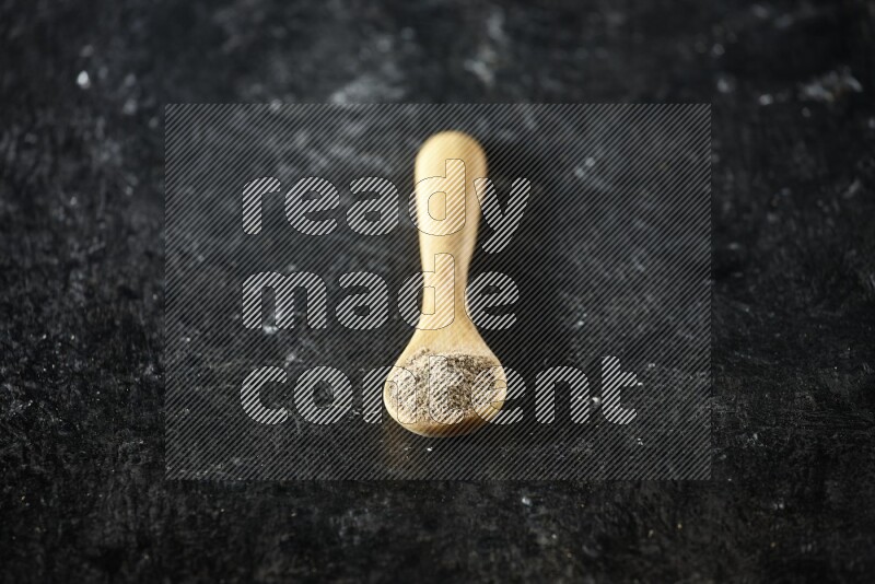 A wooden spoon full of cardamom powder on textured black flooring