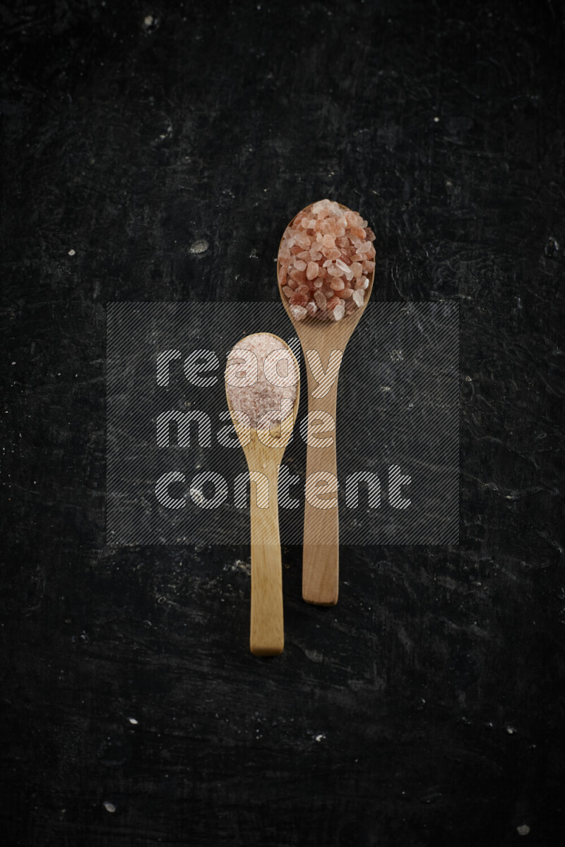 2 wooden spoons filled with fine and coarse salt on black background