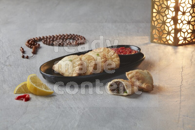 Four fried sambosas in an oval shaped black plate, beside a cut cheese sambosa, a brown misbaha and a golden lantern on a gray background
