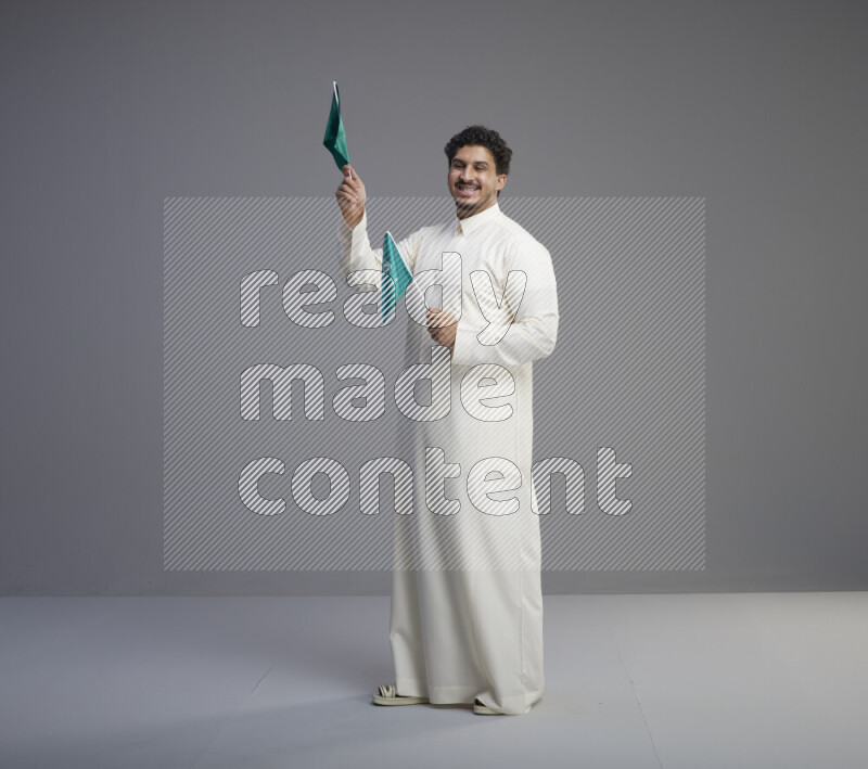 A Saudi man standing wearing thob holding small Saudi flag on gray background