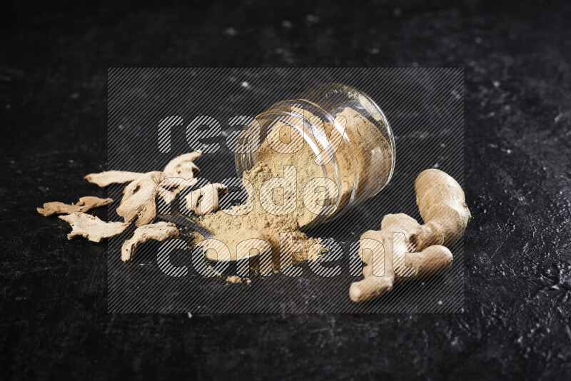 A glass jar full of ground ginger powder flipped with some spilling powder on black background