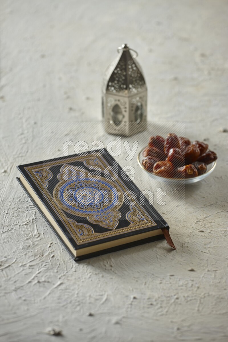 A silver lantern with different drinks, dates, nuts, prayer beads and quran on textured white background